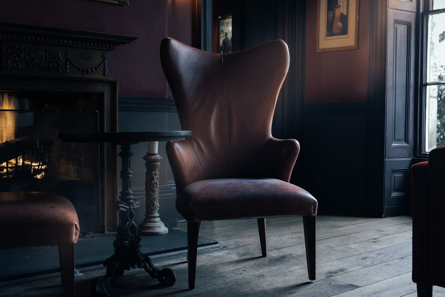 A high backed wing chair in worn dark leather standing on bare boards beside a lit fireplace in a dim panelled room.