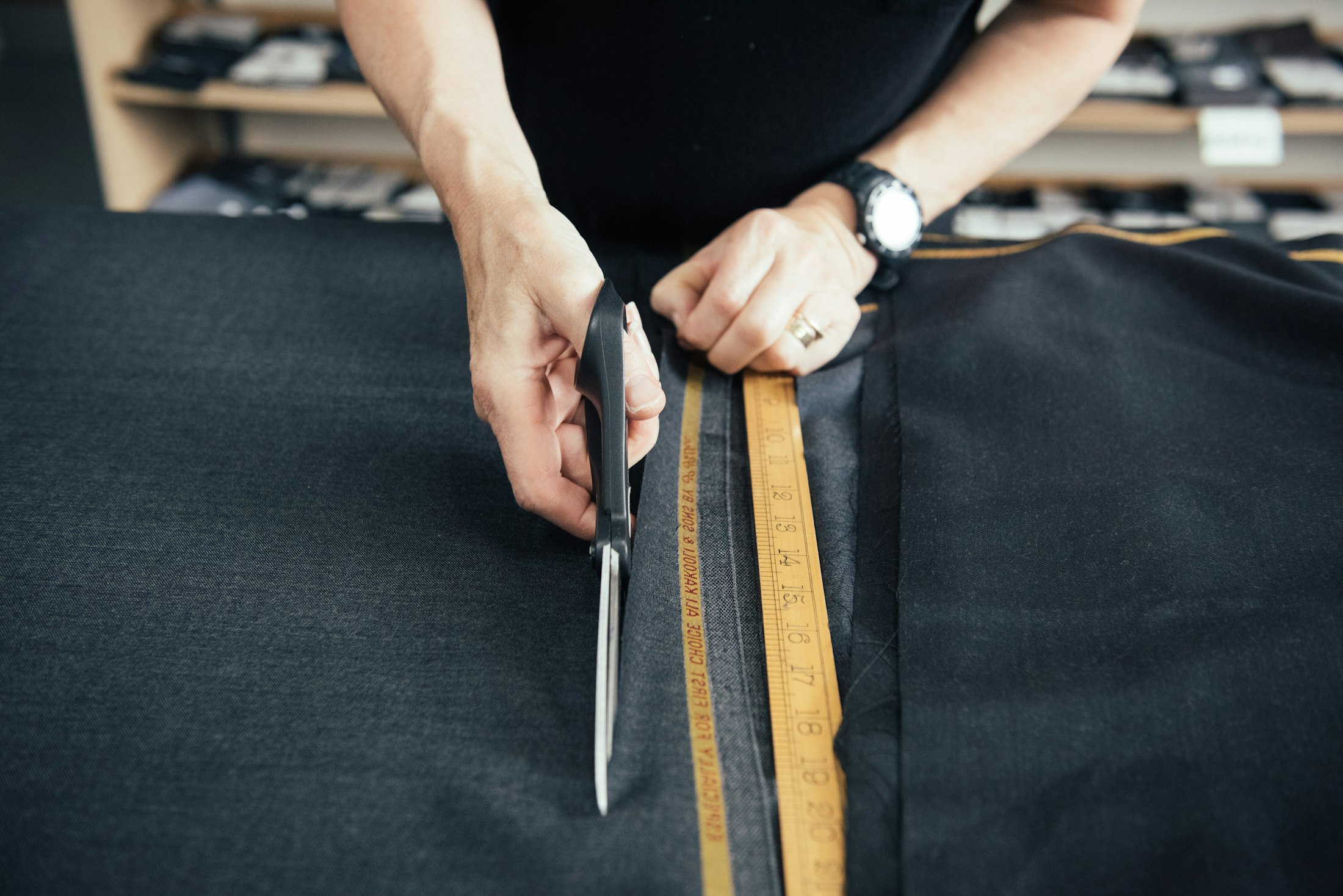 Two hands cutting into a length of dark cloth on a bench with heavy shears, a folding boxwood yardstick laid along the cut line and shelves of folded goods behind.