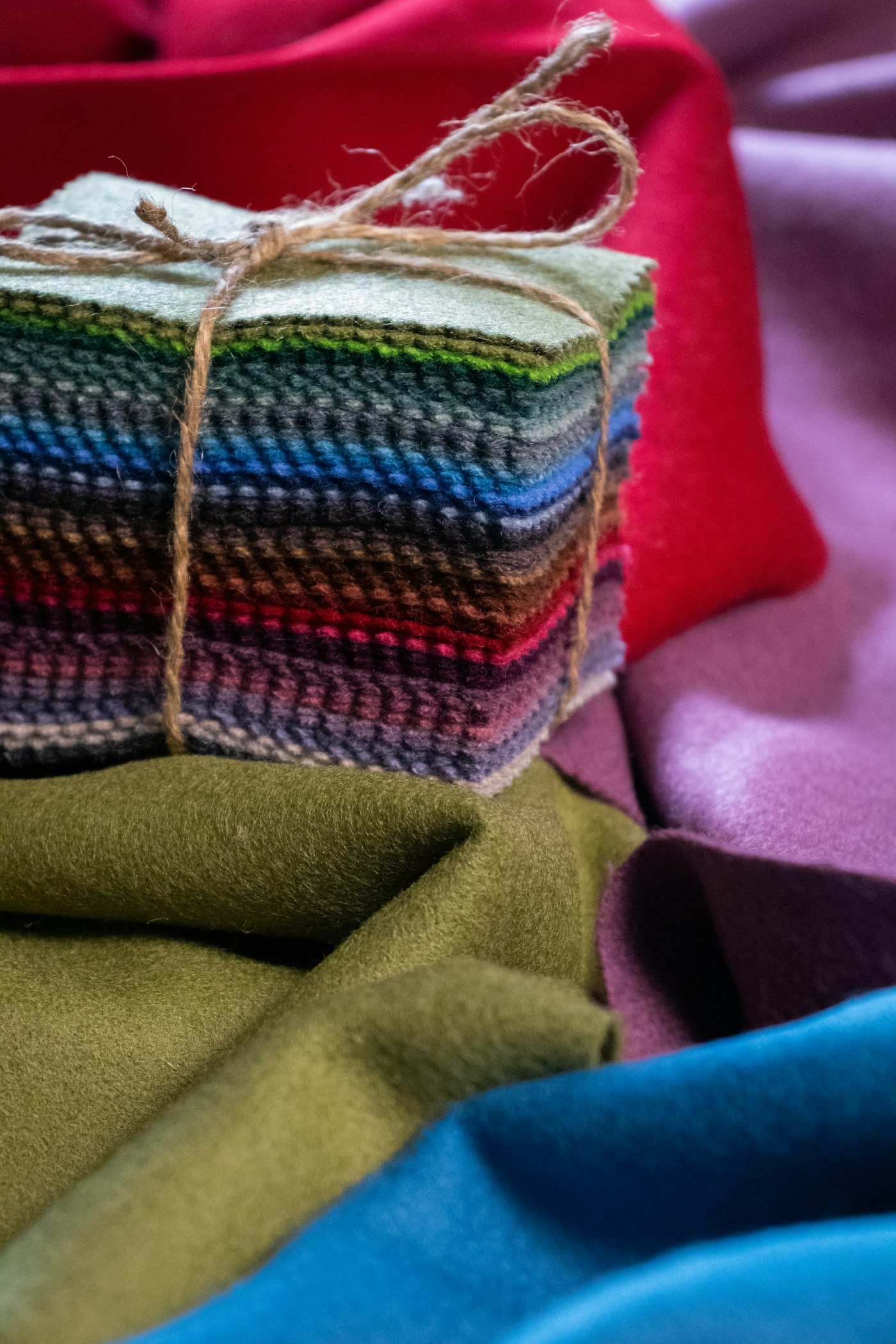 A bundle of upholstery weight wool swatches tied with garden twine, resting on folded lengths of olive, wine and teal cloth.